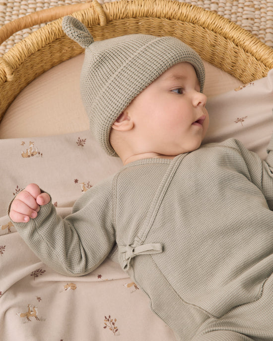 A baby lying on a patterned blanket inside a woven bassinet, wearing a textured sage green knit hat with a small pom-pom and a matching sage green long-sleeve romper.