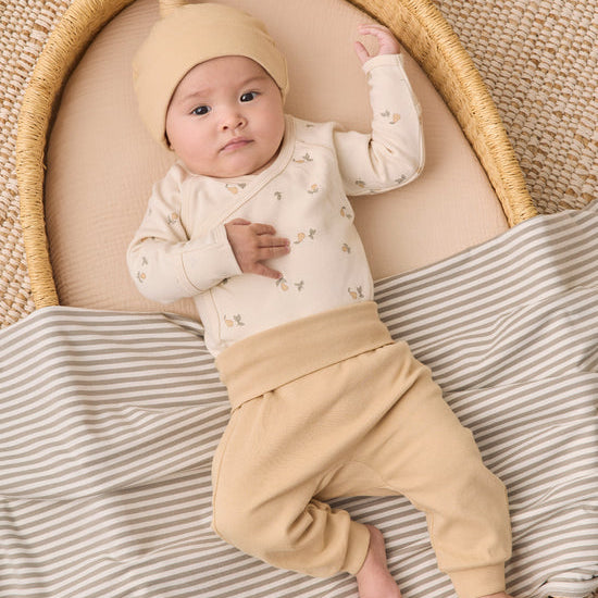 A baby wearing a cream long-sleeve top with tiny floral prints, tan pants, and a matching beanie hat, lying on a striped blanket inside a woven bassinet.