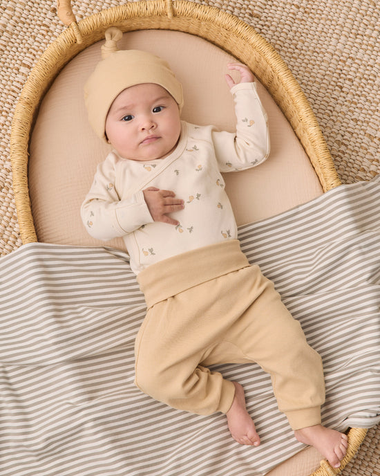 A baby wearing a cream long-sleeve top with tiny floral prints, tan pants, and a matching beanie hat, lying on a striped blanket inside a woven bassinet.