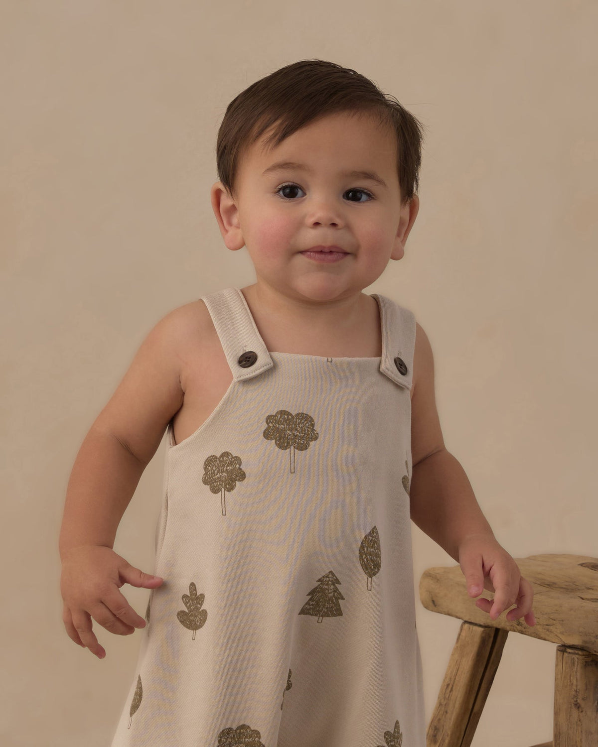 A toddler wearing a sleeveless cream-colored romper with a metallic gold tree pattern, standing next to a rustic wooden stool against a warm beige background.