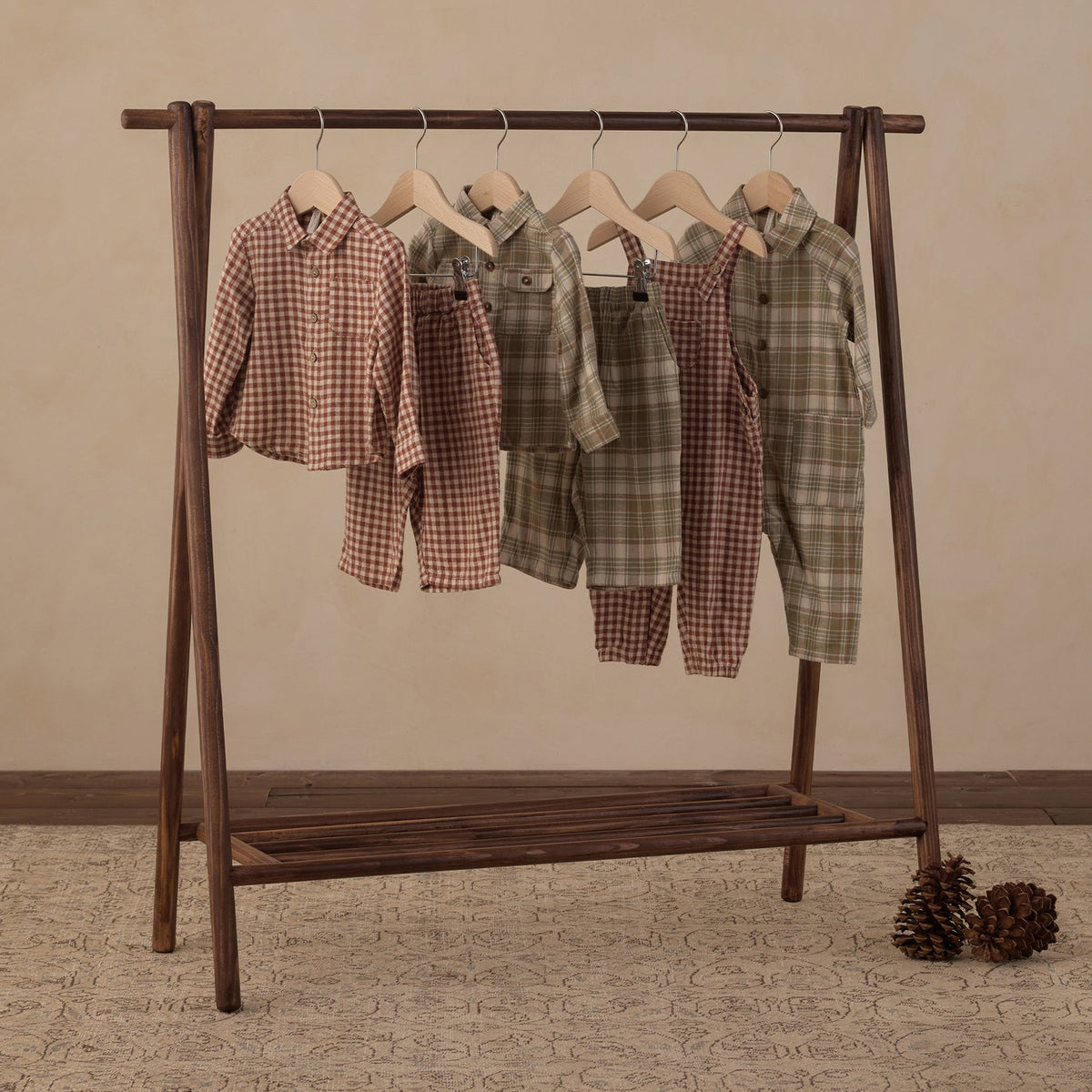 Five children's two-piece pajama sets in plaid and gingham patterns hang on a dark wooden A-frame clothing rack. The sets feature earthy tones including brown, green, and beige.