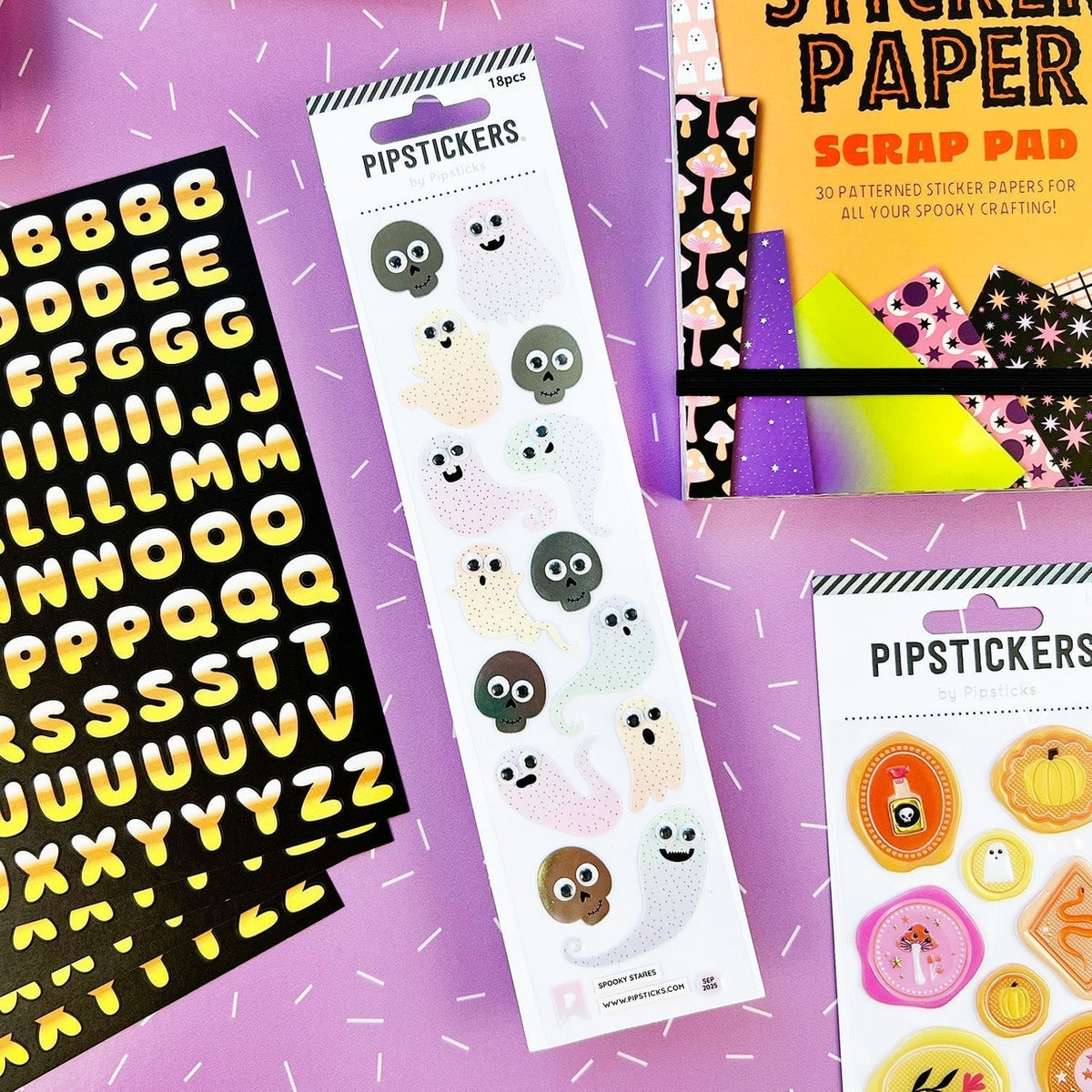 A styled flat-lay of Halloween-themed sticker products on a purple background. The products include a sheet of black alphabet stickers with yellow letters, a sheet of white ghost stickers with colorful faces, and a sheet of autumn pumpkin and face stickers.