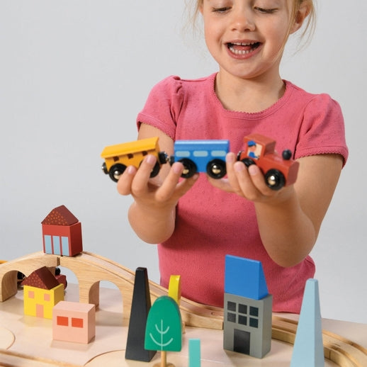 A young child with blonde hair wearing a pink shirt holds up a blue toy train engine and yellow cargo car, smiling playfully above a wooden toy train set and colorful town blocks.