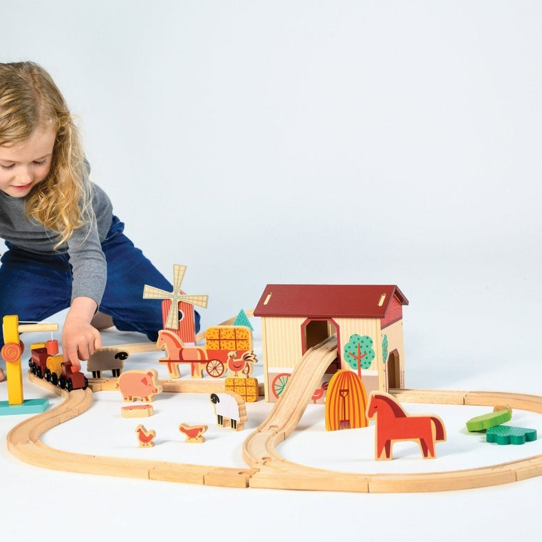 A young child lying on the floor, reaching out to play with a wooden farmyard-themed train set complete with tracks, a red barn, a windmill, and various animal figures.