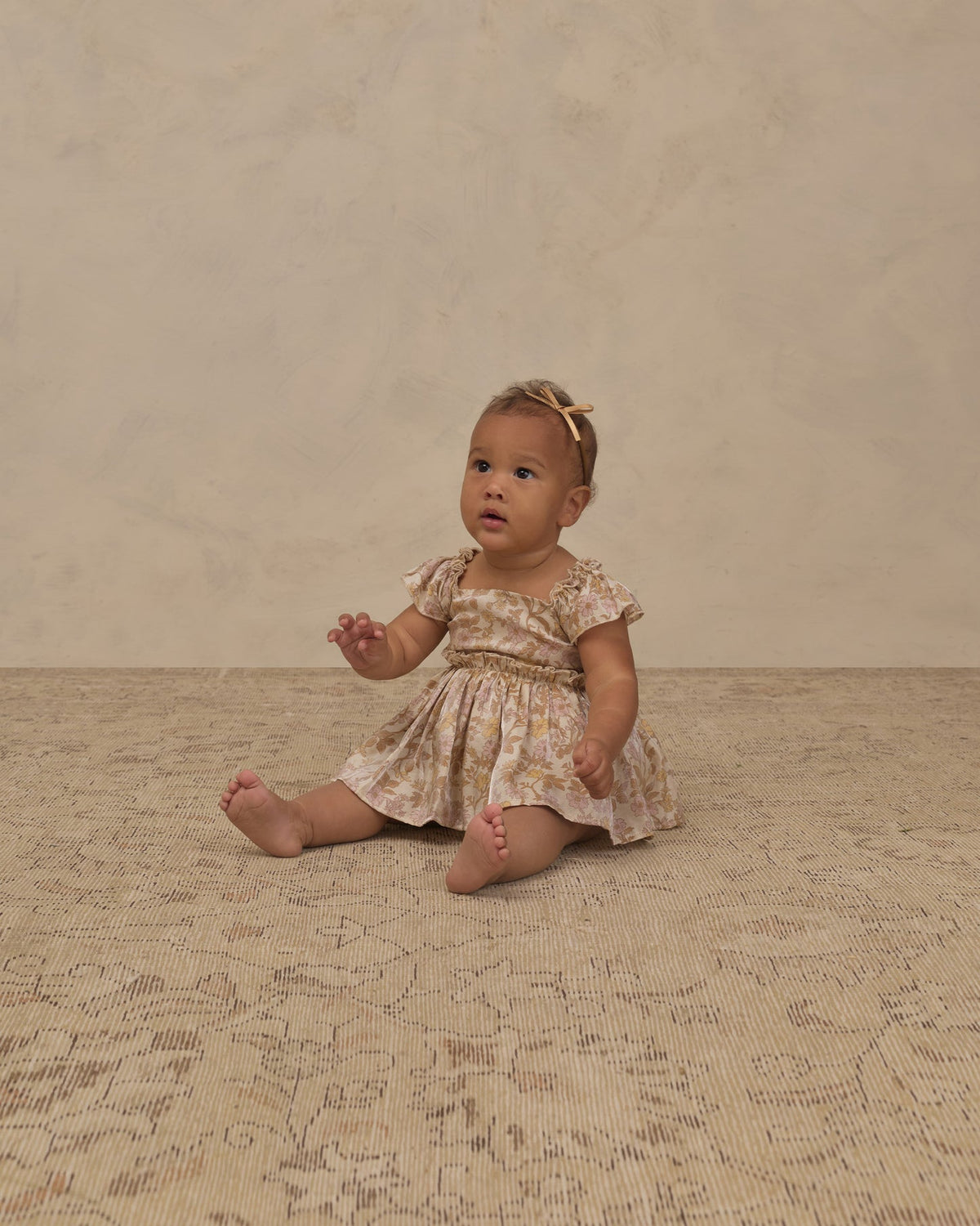 A baby girl sitting on a textured beige rug in front of a neutral beige backdrop, wearing a short-sleeve cream dress with a subtle floral pattern and a matching hair bow.