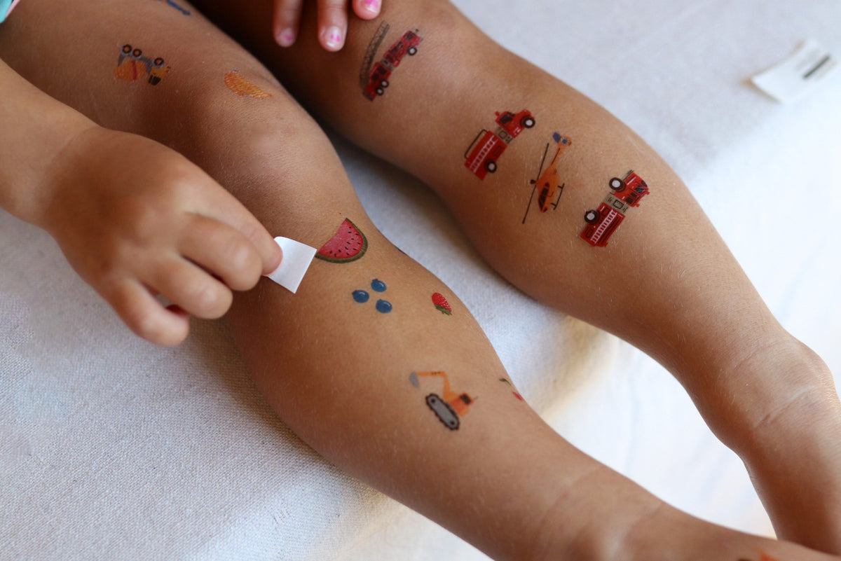 Children's legs with temporary tattoos of cars and a watermelon, being cleaned by a hand.
