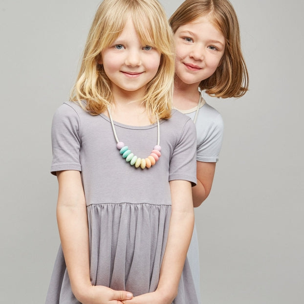 Two young girls modeling short-sleeve tiered dresses in a studio setting. The girl in the foreground wears a dark charcoal gray dress and poses with her arms crossed, while the girl behind her wears a light gray dress.