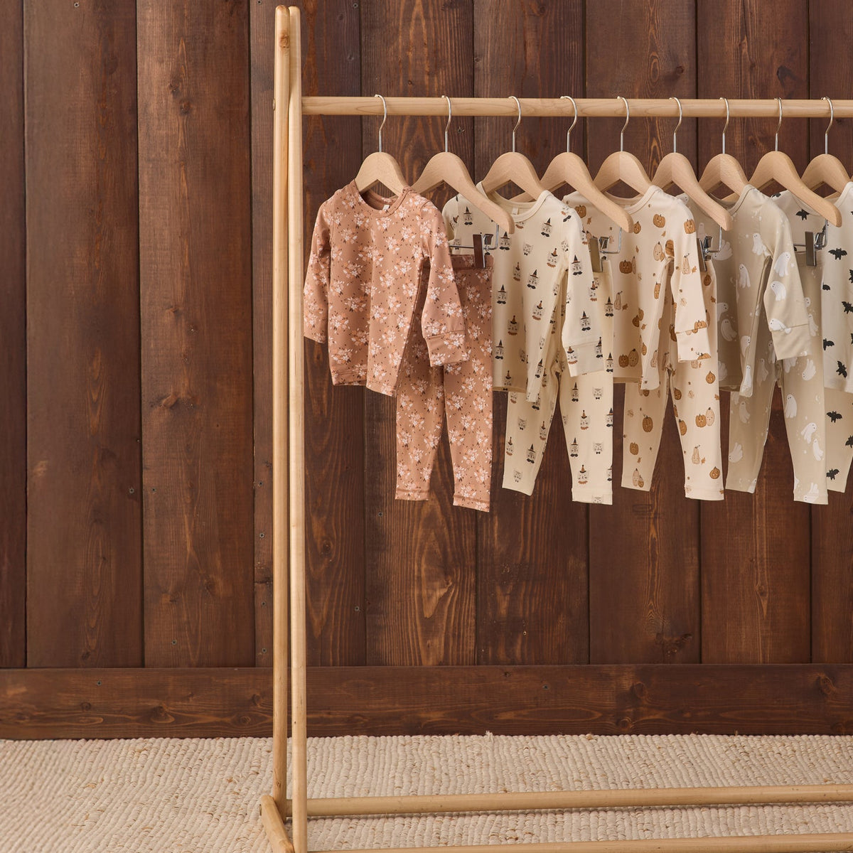 A wooden clothing rack holding several baby long-sleeve one-piece outfits in soft neutral tones, displayed against a dark wood plank wall and light woven rug.