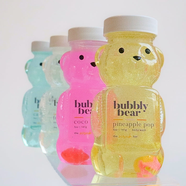 Four translucent bear-shaped body wash bottles lined up on a white background, with the front bottle labeled 'bubbly bear pineapple pop' in yellow glittery liquid and the others in pastel blue, pink, and white.