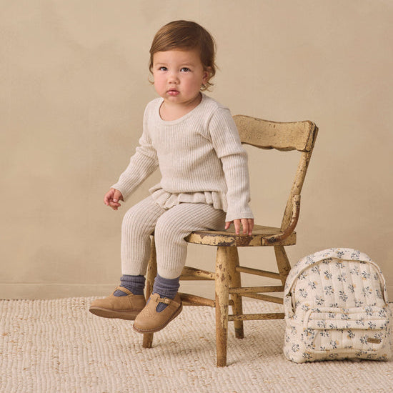A toddler sits on a rustic wooden chair wearing a cream ribbed knit sweater and matching pants, with blue socks and tan shoes. A small floral-patterned backpack rests beside the chair against a warm beige studio backdrop.