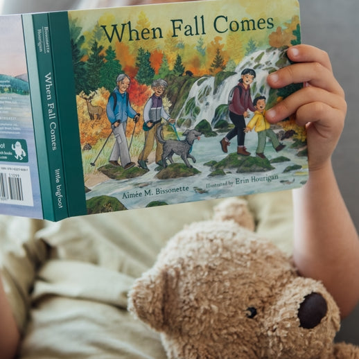 A toddler holding an open board book titled When Fall Comes, looking down at the colorful illustrated pages showing autumn trees and an outdoor nature scene, with a teddy bear resting nearby.