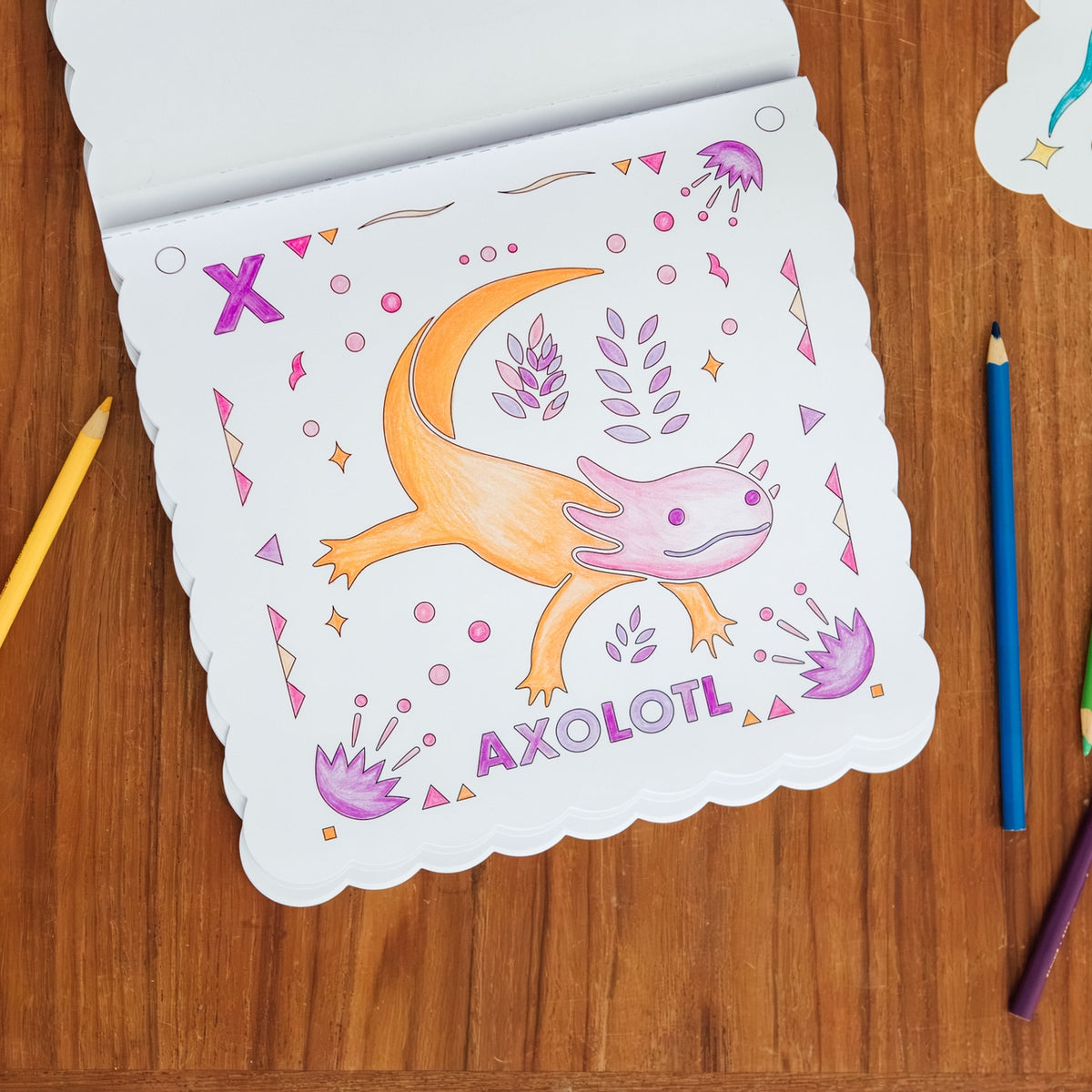 A scalloped-edge coloring page featuring a cartoon axolotl with the word 'AXOLOTL' printed below, placed on a wooden surface with colored pencils around it.