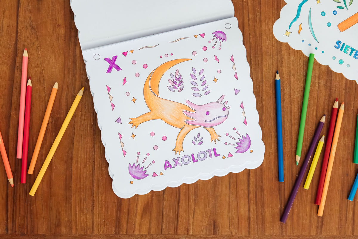 A scalloped-edge coloring page featuring a cartoon axolotl with the word 'AXOLOTL' printed below, placed on a wooden surface with colored pencils around it.