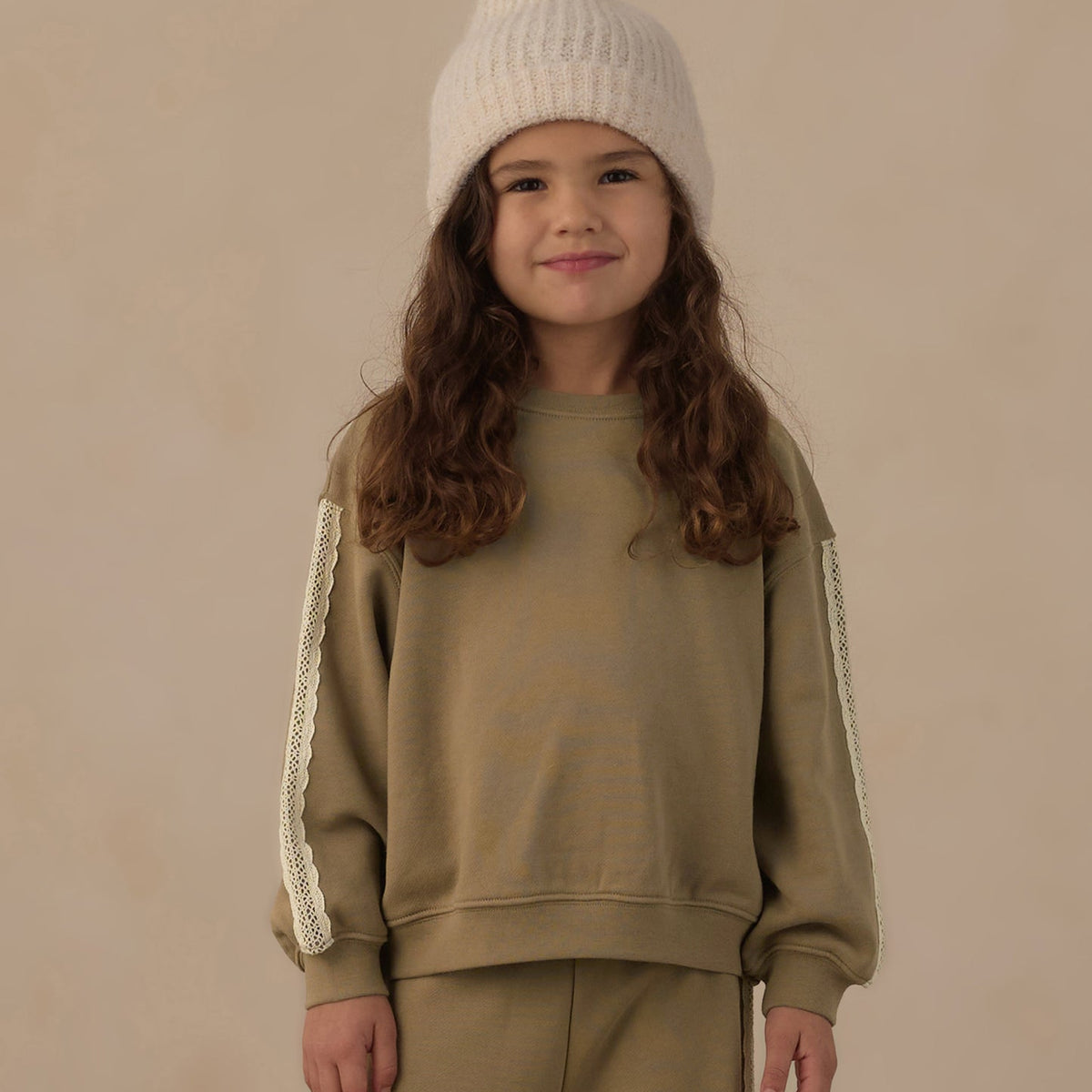 A young girl with long wavy brown hair is wearing a matching beige sweatshirt and sweatpants set with cream lace sleeve details, paired with a cream knit beanie, against a solid beige background.