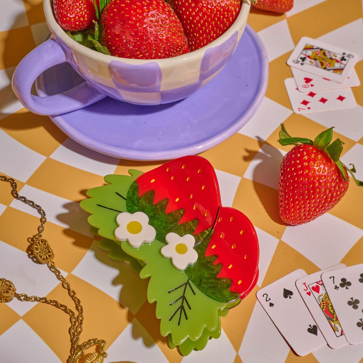 Strawberries in a teacup on a checkered tablecloth with playing cards and a necklace.