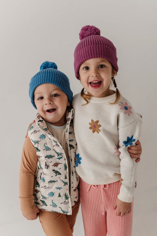 Two young children smiling and posing together against a plain light gray studio background. The child on the left wears a blue knit beanie, a cream vest with a dinosaur print, and tan pants.