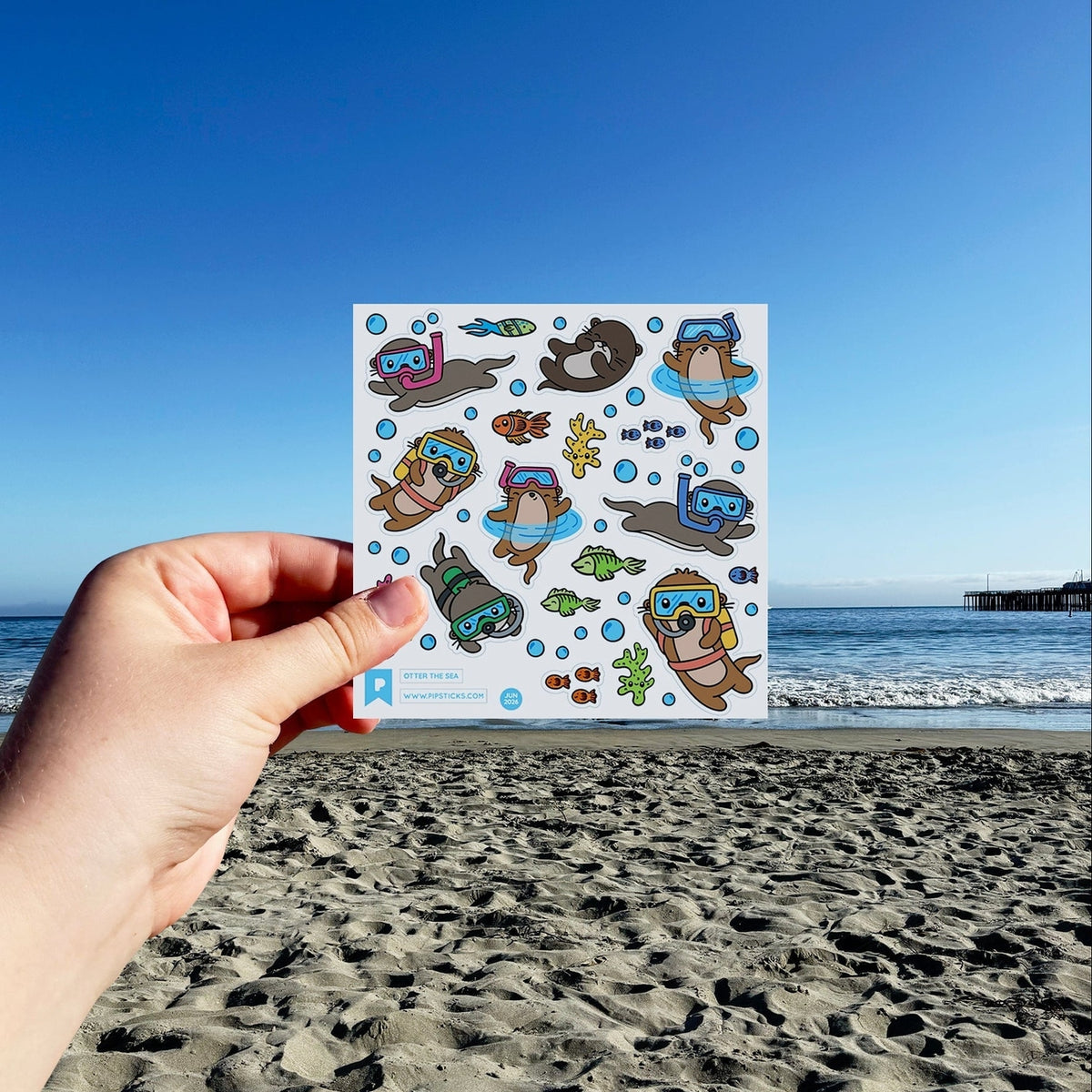 A hand holds a square sticker sheet featuring cartoon sea otters and fish in swimwear and diving gear, set against a real beach and ocean background under a clear blue sky.