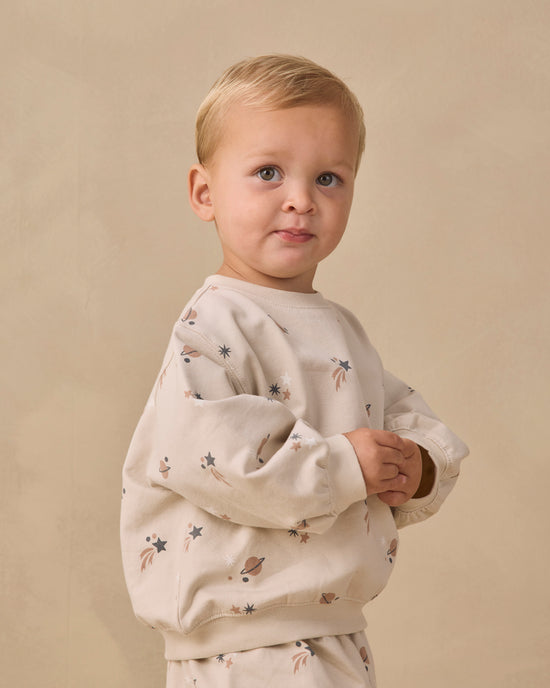 A young toddler with blonde hair is wearing a cream-colored crewneck sweatshirt and matching pants set, both decorated with a small celestial print of stars, planets, and rockets. The child is standing against a warm, textured beige studio backdrop.