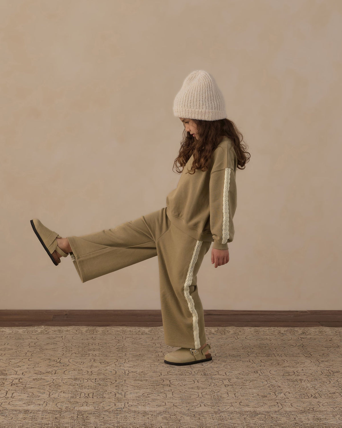 A child wearing a matching beige tracksuit with white side stripes, a cream knit beanie, and tan shoes, posing with one leg raised against a textured beige wall and patterned rug.