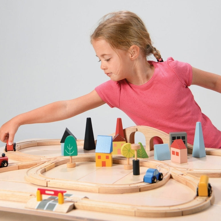 A young child in a pink shirt sits behind a wooden train set laid out on a white surface, reaching across to place a red toy car onto the tracks. The setup features a figure-eight track configuration with a blue train engine, cargo cars, and colorful wooden town elements like houses, trees, and a bridge.