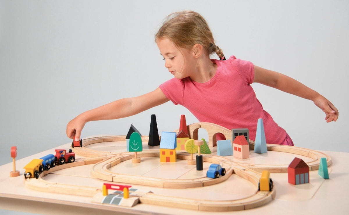 A young child in a pink shirt sits behind a wooden train set laid out on a white surface, reaching across to place a red toy car onto the tracks. The setup features a figure-eight track configuration with a blue train engine, cargo cars, and colorful wooden town elements like houses, trees, and a bridge.