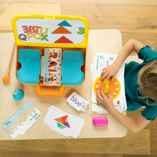 Child playing with educational toys and materials on a wooden table, including a colorful box labeled 'EBBTU QPOX'.