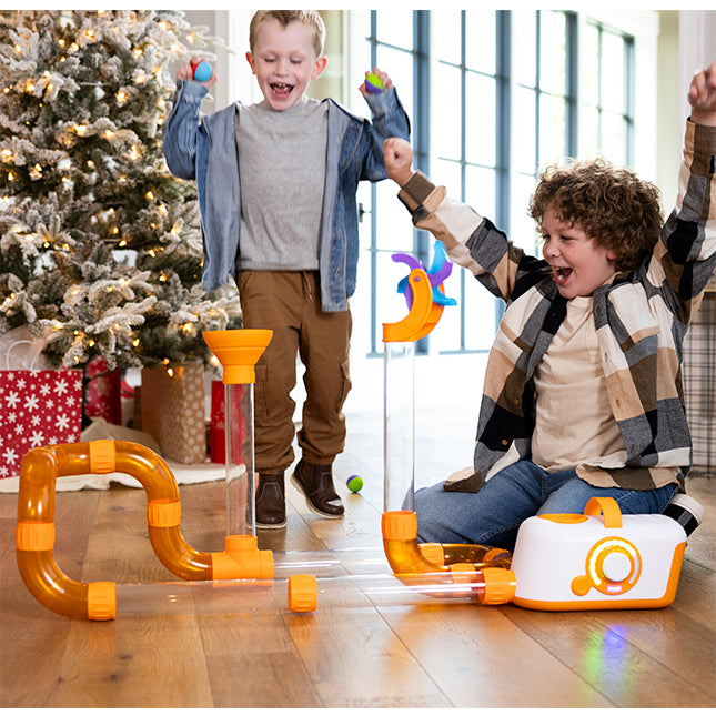 Two children playing with a toy set on a wooden floor with a Christmas tree in the background.