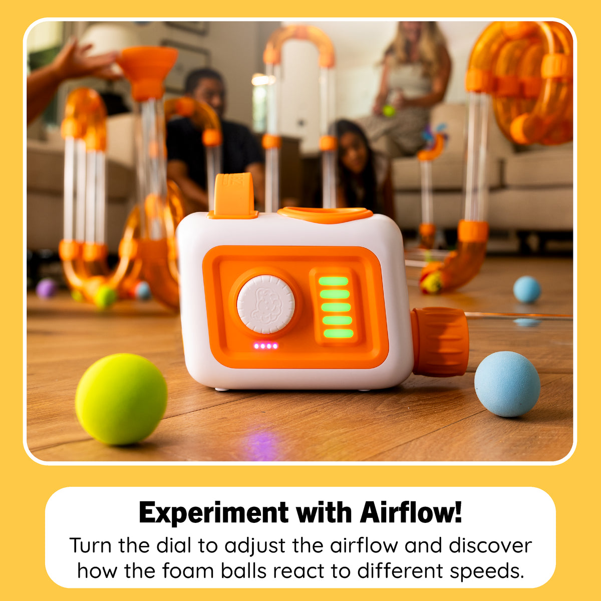 A white and orange robotic toy with a dial and LED lights sits on a wooden floor in a living room, with people blurred in the background playing with similar foam balls and tubing. Text at the bottom reads, 'Experiment with Airflow! Turn the dial to adjust the airflow and discover how the foam balls react to different speeds.