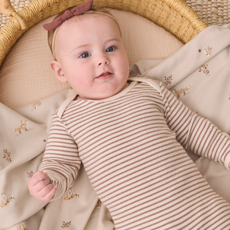 A baby lying in a woven bassinet, wearing a beige and brown striped long-sleeve bodysuit with a dusty rose bow headband.