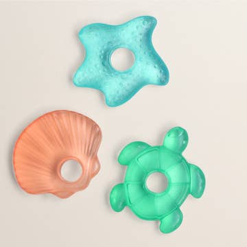 Three colorful baby teething rings shaped like a starfish, shell, and turtle on a light beige background.