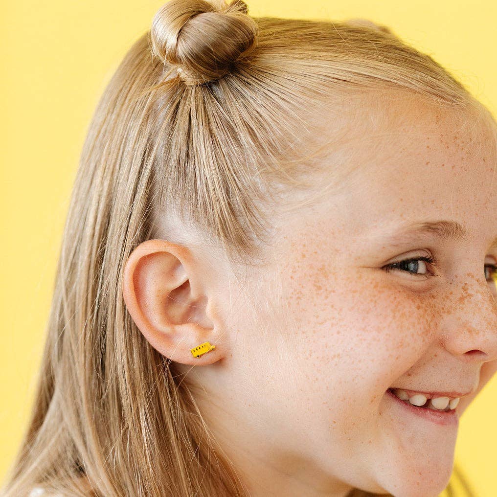 Close-up of a girl with blonde hair tied in a bun, wearing a yellow earring against a yellow background.