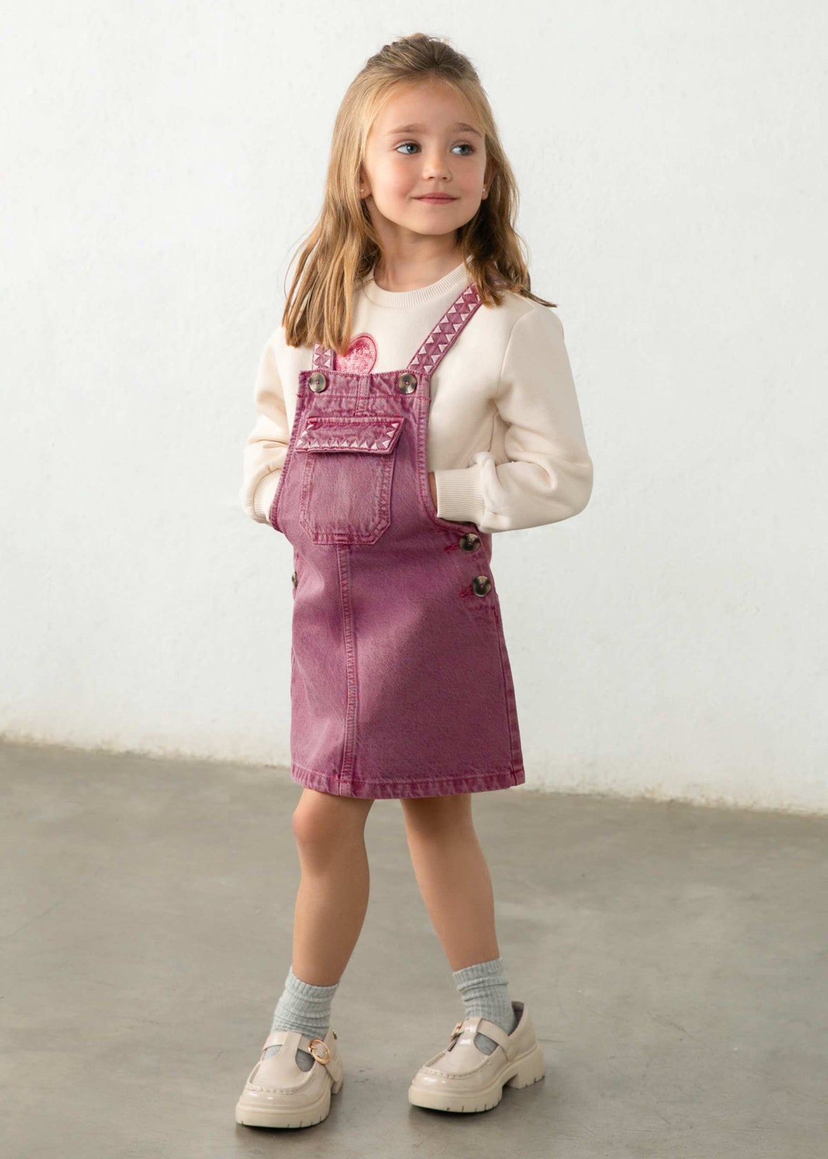 A child wearing dusty rose denim overall with pink embroidered details and a cream sweatshirt underneath, shown from the chest up against a light neutral background.