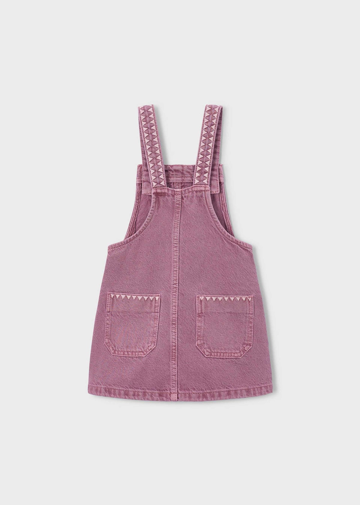 A dusty pink denim overall dress for kids, featuring adjustable shoulder straps with a zigzag pattern, side pockets with metal snap buttons, and a front bib pocket, shown against a plain white background.