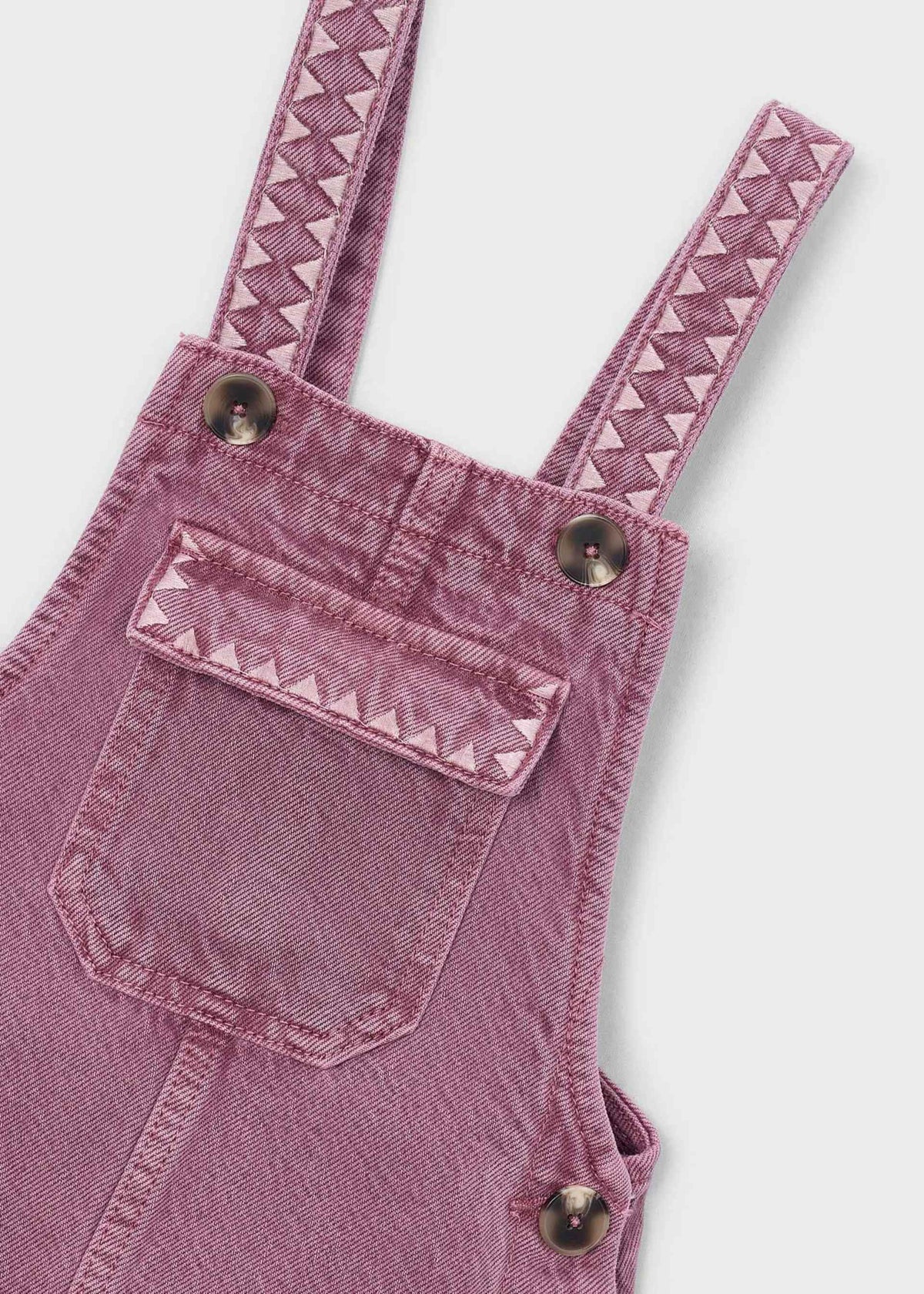 A dusty pink denim overall dress for kids, featuring adjustable shoulder straps with a zigzag pattern, side pockets with metal snap buttons, and a front bib pocket, shown against a plain white background.