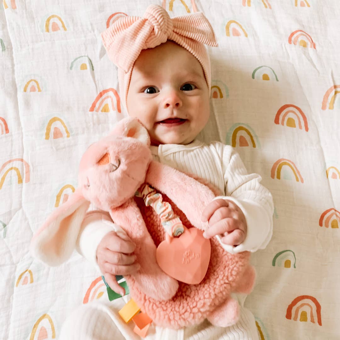 Baby holding a pink plush toy with a rainbow-patterned blanket in the background