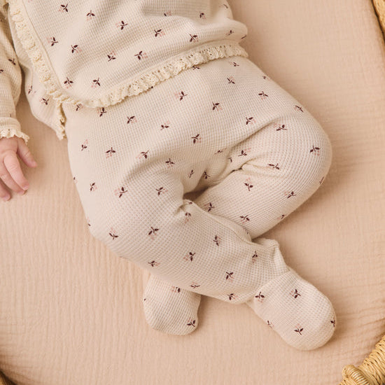 A baby wearing a cream-colored waffle-knit footed sleeper with a small brown floral pattern and lace trim, lying on a beige muslin blanket inside a woven basket.