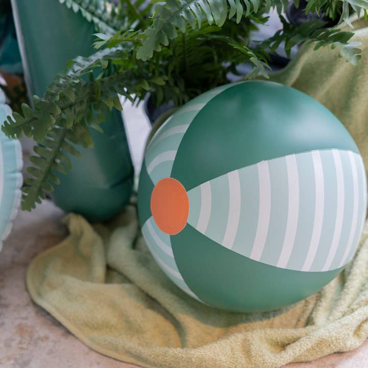 A green and light blue striped beach ball by Quut Toys featuring an orange center circle, resting on a textured cloth surface with green foliage in the background.