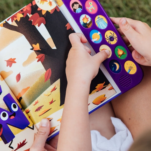 A child holding open "What Does Ñ Sound Like?: A Spanish-English Sound Book," showing an interior illustrated page with a purple animated character resembling the letter Ñ, set against an autumn landscape with falling leaves.