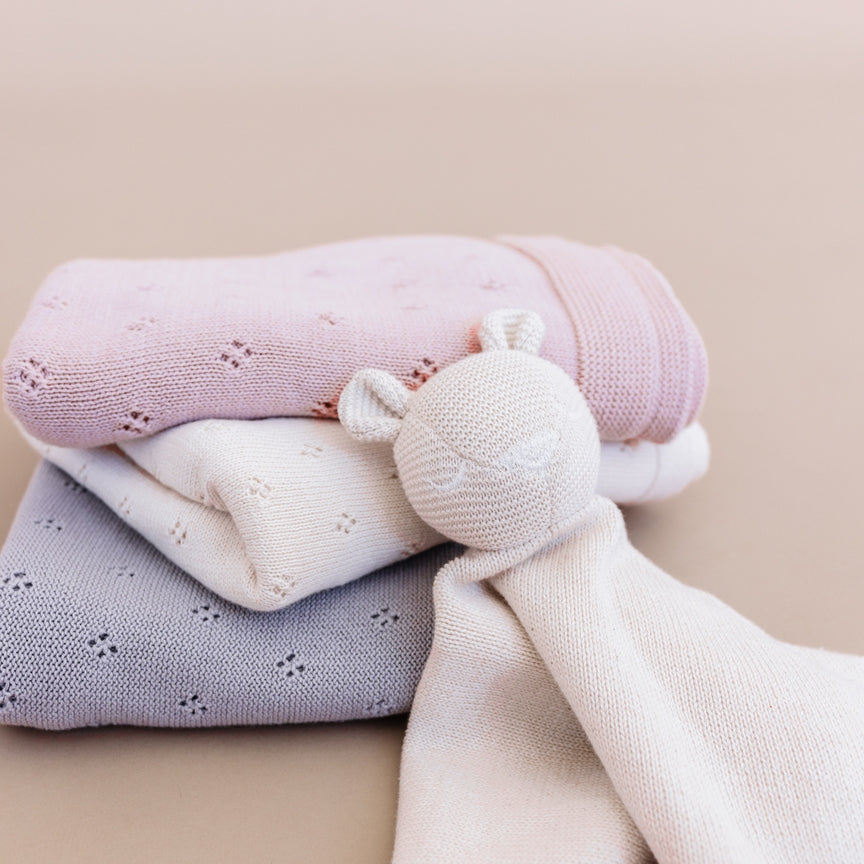 Three folded knitted baby security blankets in pastel pink, white, and light lavender-gray, with a cream knitted bear-shaped teether attached, on a soft beige background.