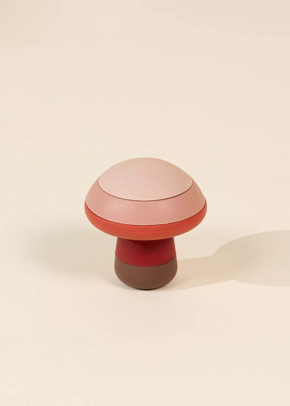 A mushroom-shaped container or candle with a light pink domed lid, a terracotta red band, a dark red middle section, and a brown base, set against a warm off-white background.