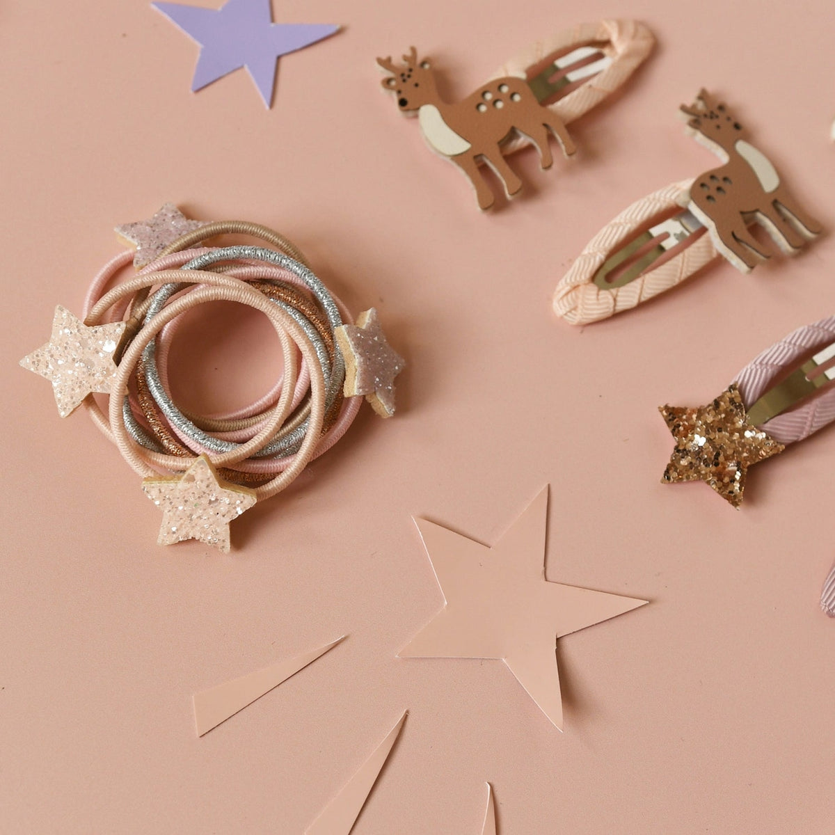 A styled flat-lay of hair accessories on a dusty pink background, featuring a bundle of pastel elastic hair ties with glittery star accents, two deer-shaped hair clips, and two star-shaped hair clips.