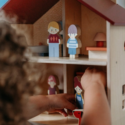 A young child playing with an open-faced wooden dollhouse by Mentari, arranged on a white table with miniature wooden furniture and small figurines.
