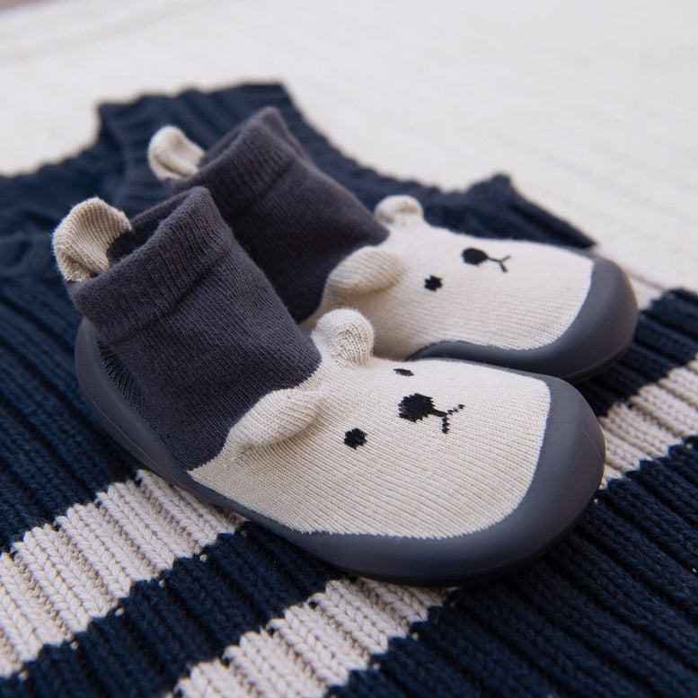 A pair of baby slip-on shoes with a bear face design, featuring a dark gray knit sock upper and a cream-colored toe section with embroidered black facial details, resting on a navy and cream striped knit blanket.