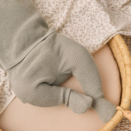 A close-up of a baby's foot wearing a sage green waffle-knit bootie, resting on a beige mattress inside a woven rattan bassinet with a floral blanket.