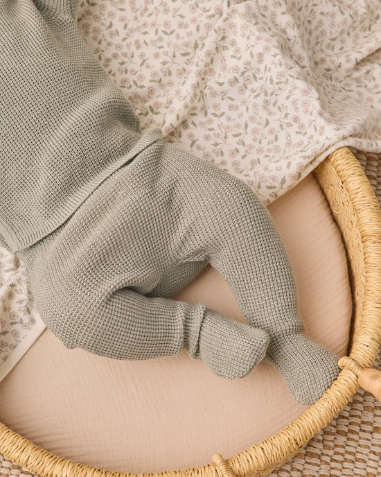 A close-up of a baby's foot wearing a sage green waffle-knit bootie, resting on a beige mattress inside a woven rattan bassinet with a floral blanket.