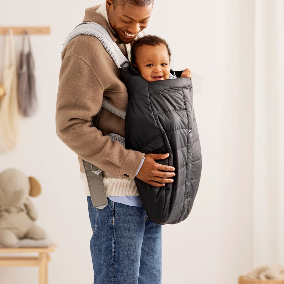 A man wearing a beige hoodie and blue jeans carries a baby in a black quilted baby carrier wrap. The scene is set in a bright, modern nursery with soft white walls, a wooden bench with a stuffed toy, and a woven basket on the floor.