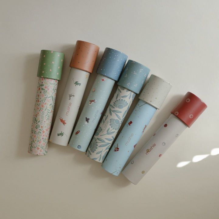 A flat lay arrangement featuring six Mushie paper kaleidoscopes in various patterns and colors—including floral, space, and dinosaur designs—lying diagonally against a neutral background with warm sunlight streaming across the lower right.