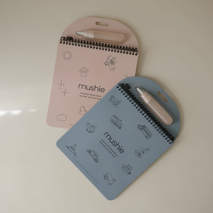 Two spiral-bound notebooks with pen holders, one pink and one blue, on a light beige background.