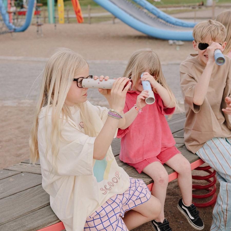 Three children sitting outdoors on a wooden bench, looking through Mushie kaleidoscopes.