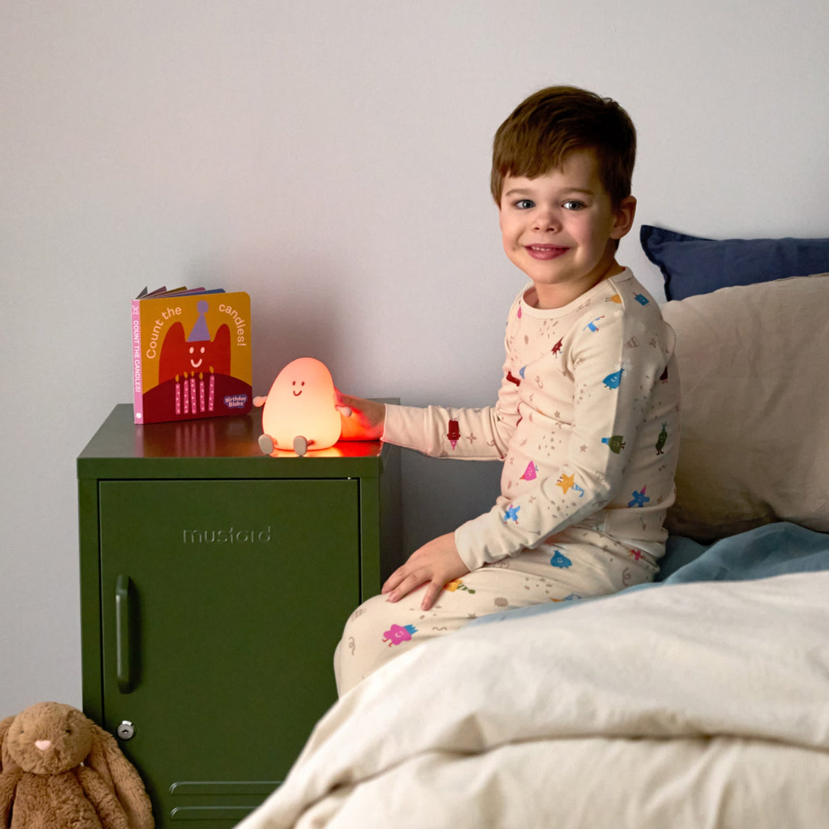 Child in pajamas sitting on a bed next to a nightstand with a night light and book